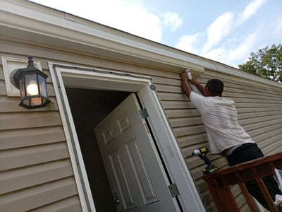 A crew member on a ladder refitting the trim above the front door, porch light on.
