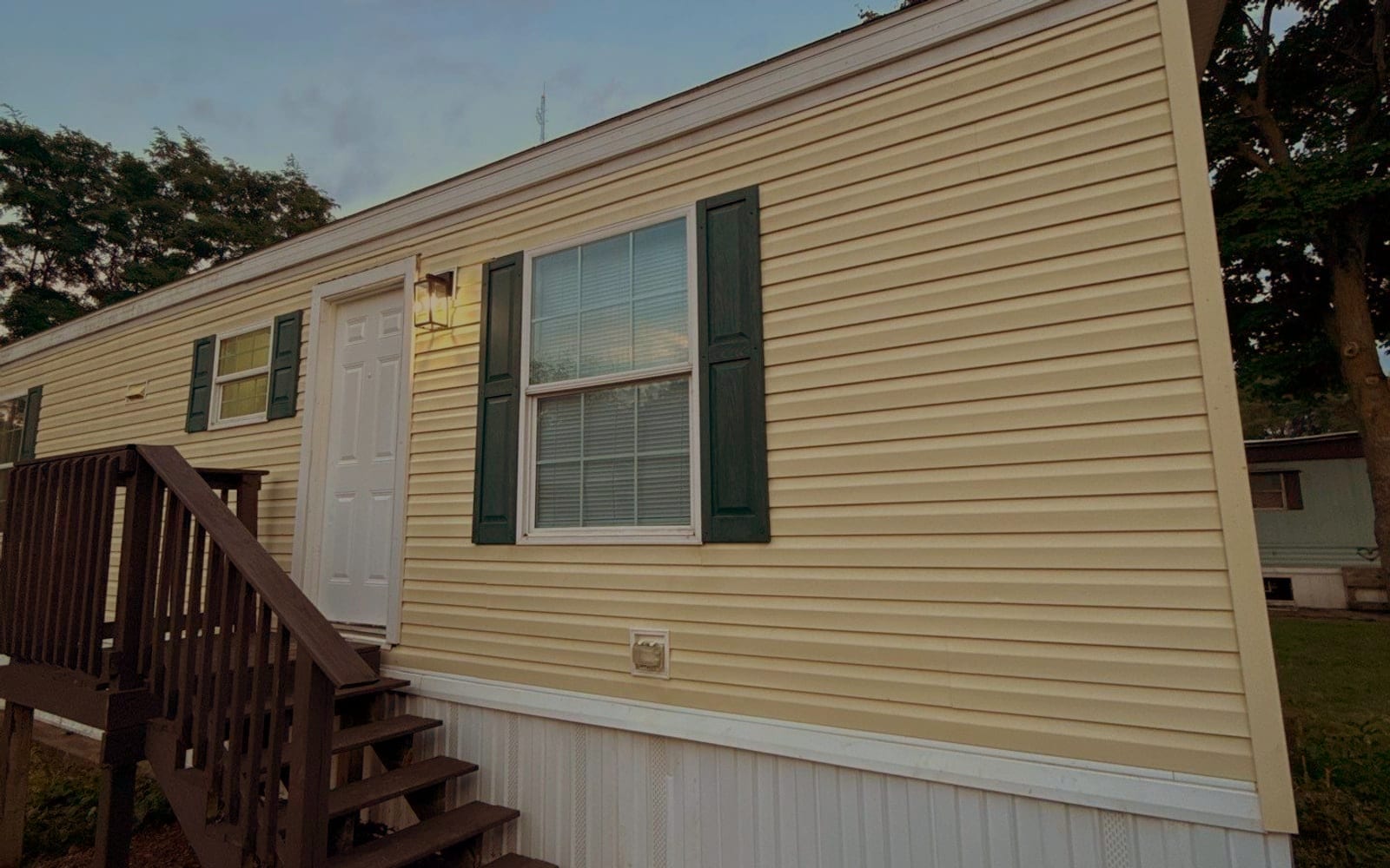 A restored single-wide at dusk - fresh cream siding, new white skirting, a rebuilt stained deck and the porch light on.